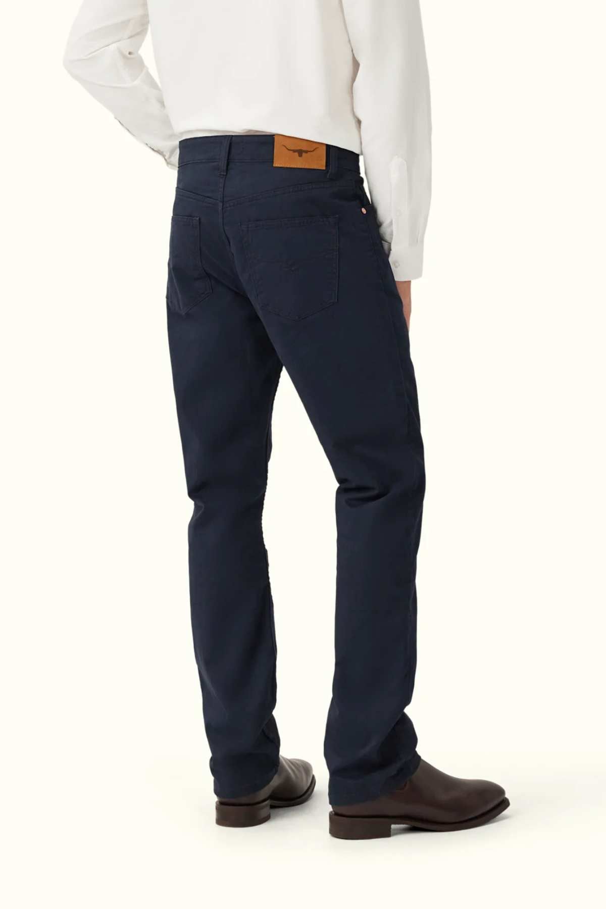 A person wearing RM Williams Ramco 5 Pocket Jeans in dark blue Australian cotton, brown boots, and a white long-sleeve shirt stands facing away, revealing the jeans’ brown leather waistband patch.