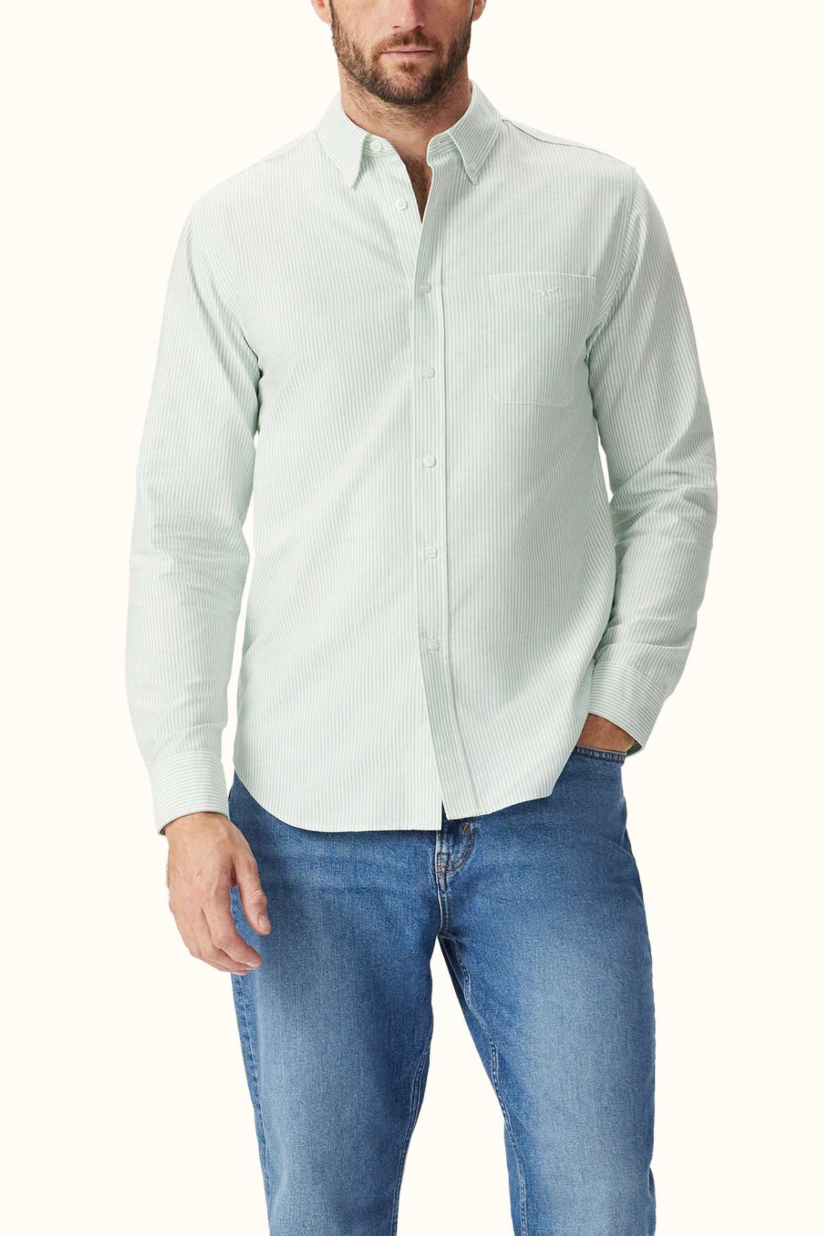 A man is wearing the RM Williams Regular Shirt in light green stripes, made from organic cotton, paired with blue jeans. This long-sleeved shirt features a pocket on the left side. He stands against a plain light background.