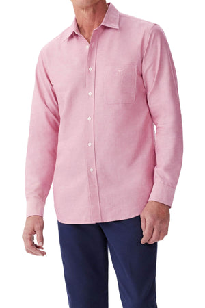 A man wears the RM Williams Regular Shirt—light pink, slim fit, long-sleeve with a collar, chest pocket, and white buttons—made from organic cotton. He pairs it with dark blue pants; his face is not visible.