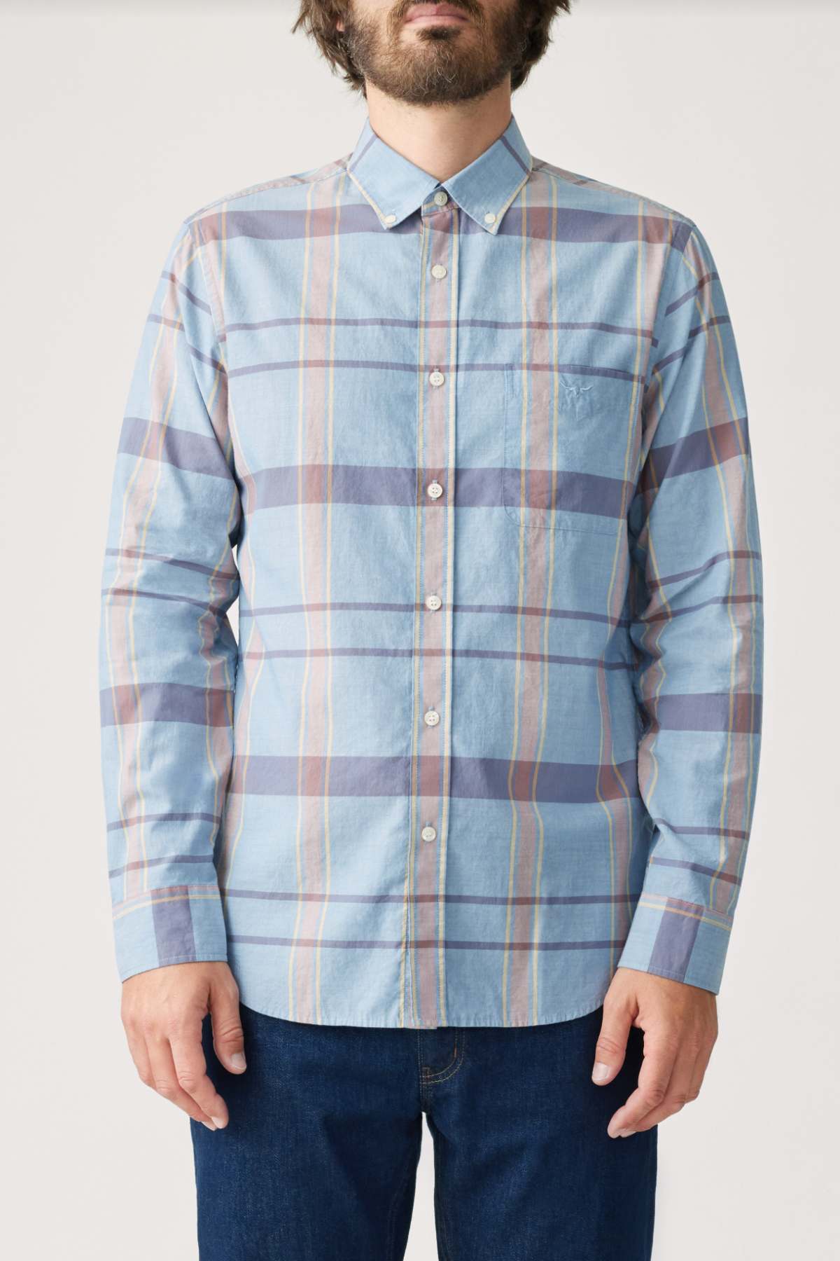 A man wears an RM Williams Regular Shirt in light blue with a multicolored plaid. The organic cotton shirt features a button-down collar, white buttons, and long sleeves. Paired with dark blue jeans, his face is partly obscured.