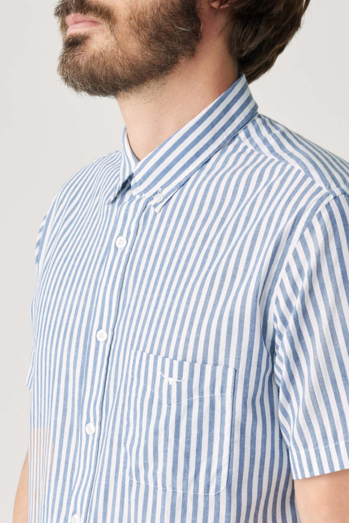 A man with a short beard and mustache wears the RM Williams Regular Short Sleeve Shirt, featuring blue and white vertical stripes, a button-down collar, chest pocket, and organic cotton. The image is cropped at forehead and mid-torso.