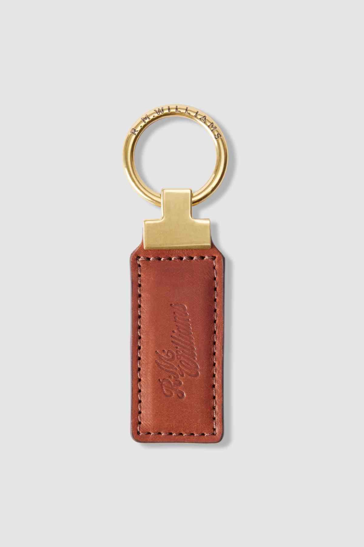 The RM Williams Ridgewood Key Fob has a rectangular brown leather strap, a gold-colored keyring, and is embossed with cursive text. This elegant and timeless design adds sophistication to key organization.