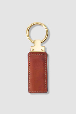 The RM Williams Ridgewood Key Fob is a brown leather rectangle with visible stitching, featuring a shiny gold round ring. It stands out against a light gray background, subtly showcasing the R.M. Williams logo.
