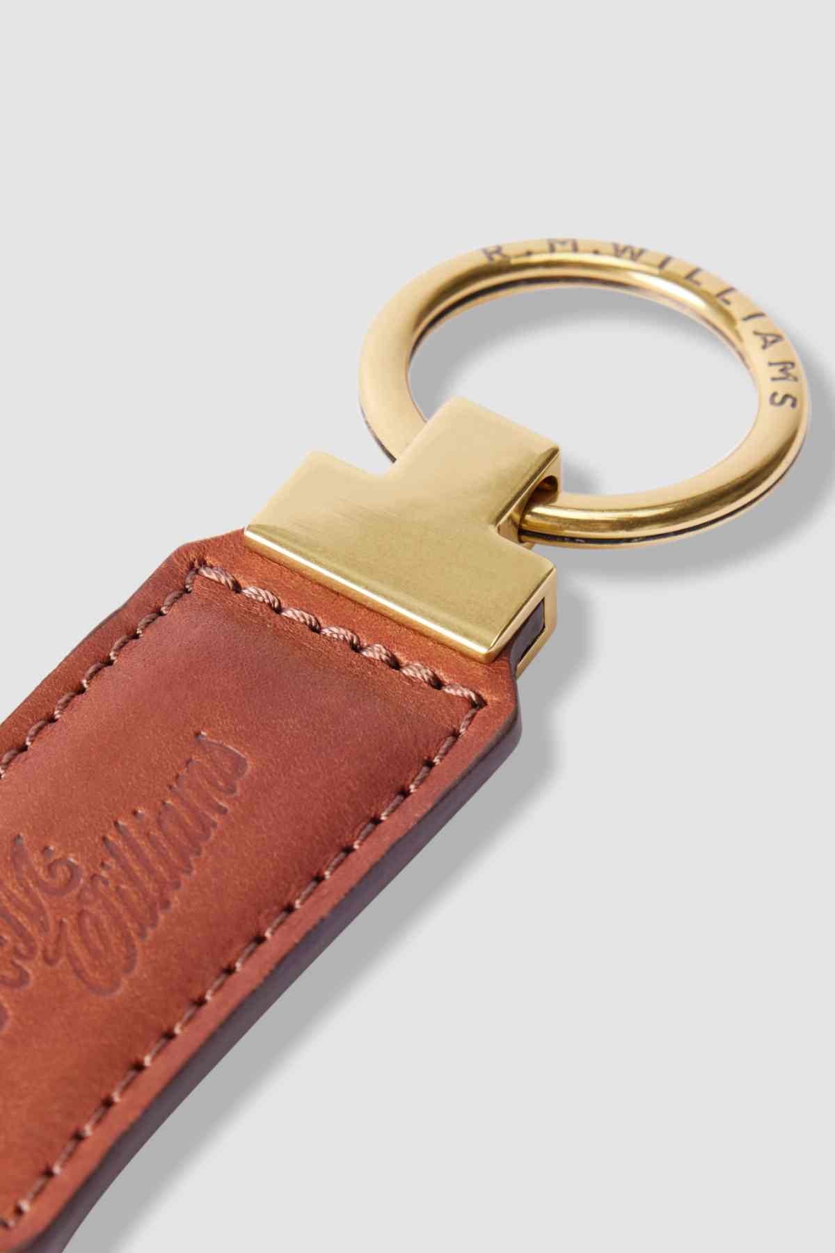 A close-up of the RM Williams Ridgewood Key Fob reveals embossed text on rich brown leather, a gold-tone metal ring with the R.M. Williams logo, and a sleek gold connector, exuding timeless sophistication.
