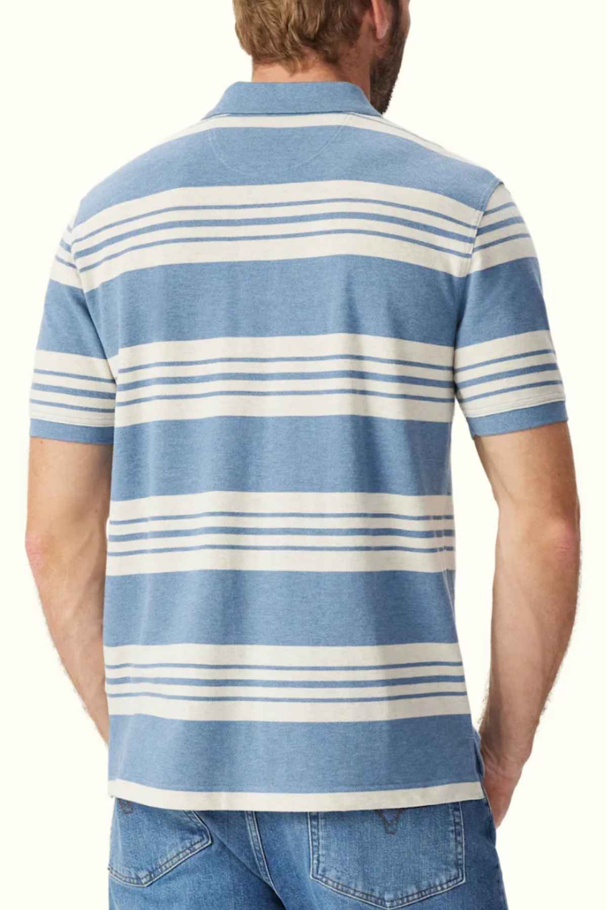 A man is standing with his back to the camera, wearing an RM Williams Rod Polo featuring short-sleeves, a collar, and blue and white horizontal stripes made from breathable cotton pique. He also has on blue jeans. His right arm is hanging by his side, and his left hand is partially tucked into his jeans pocket.