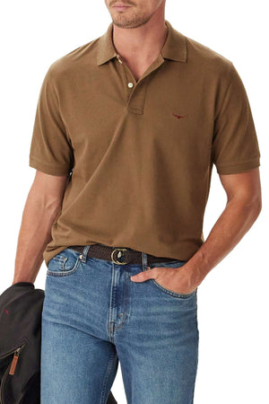 A man wears the RM Williams Rod Polo, a brown short-sleeve shirt with a small embroidered chest logo, tucked into blue jeans. He holds a jacket in one hand while his other hand rests in his pocket.