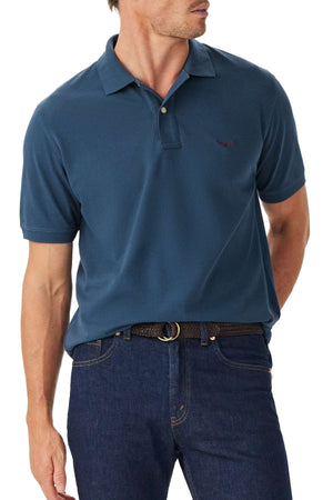 A man wearing the RM Williams Rod Polo, a short-sleeve teal shirt, tucked into dark blue jeans with a black woven belt, looking to the side.