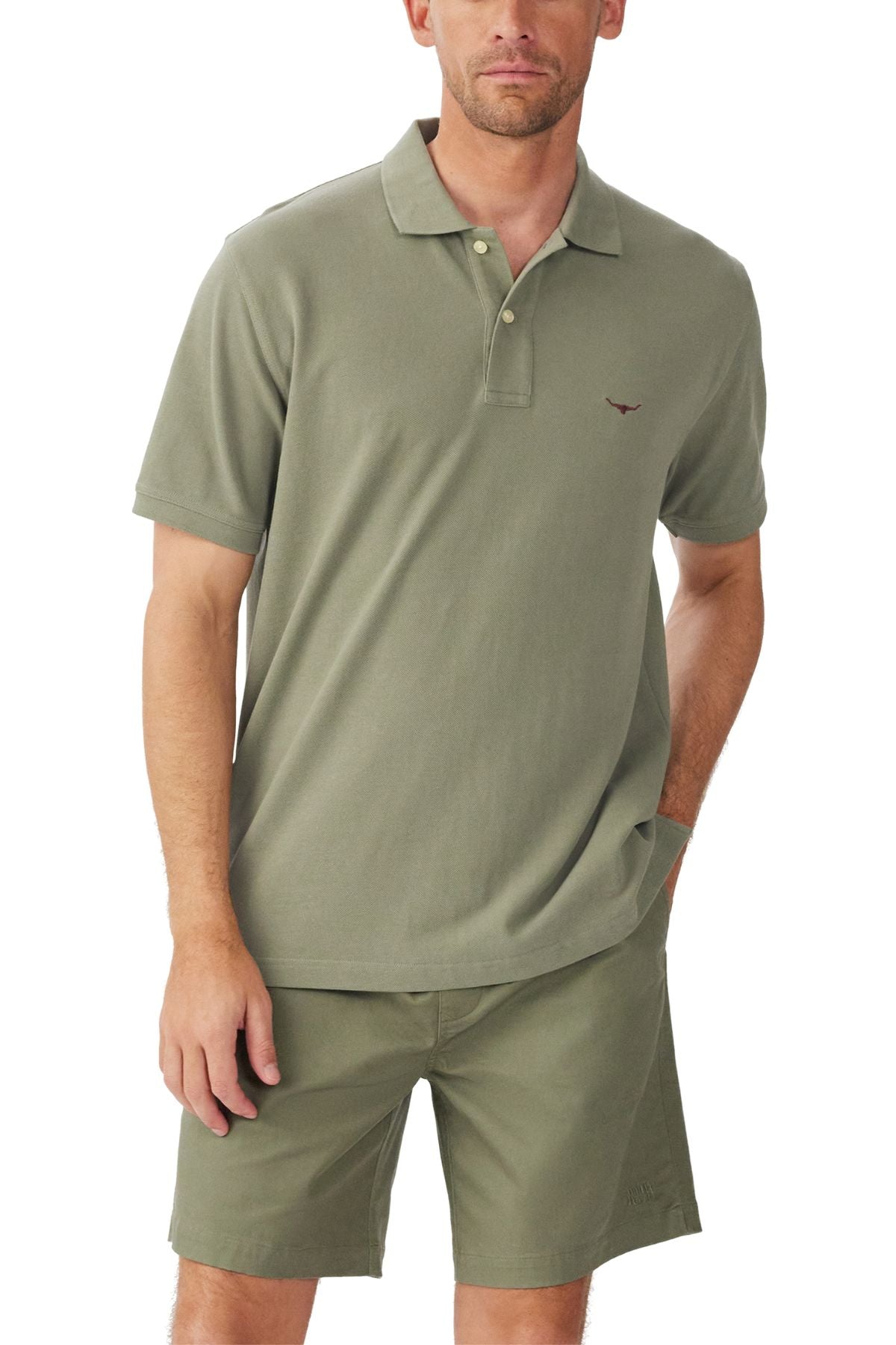 A man wears the RM Williams Rod Polo in olive green organic cotton pique with matching shorts, standing against a white background with one hand in his pocket.