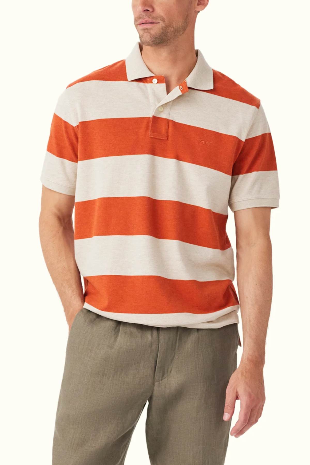 A person is wearing the RM Williams Rod Polo, featuring orange and cream stripes made from organic cotton, paired with olive green pants. Their right hand is tucked into their pocket as they look slightly to the side, highlighting a classic fit that reflects R.M.Williams' timeless style.
