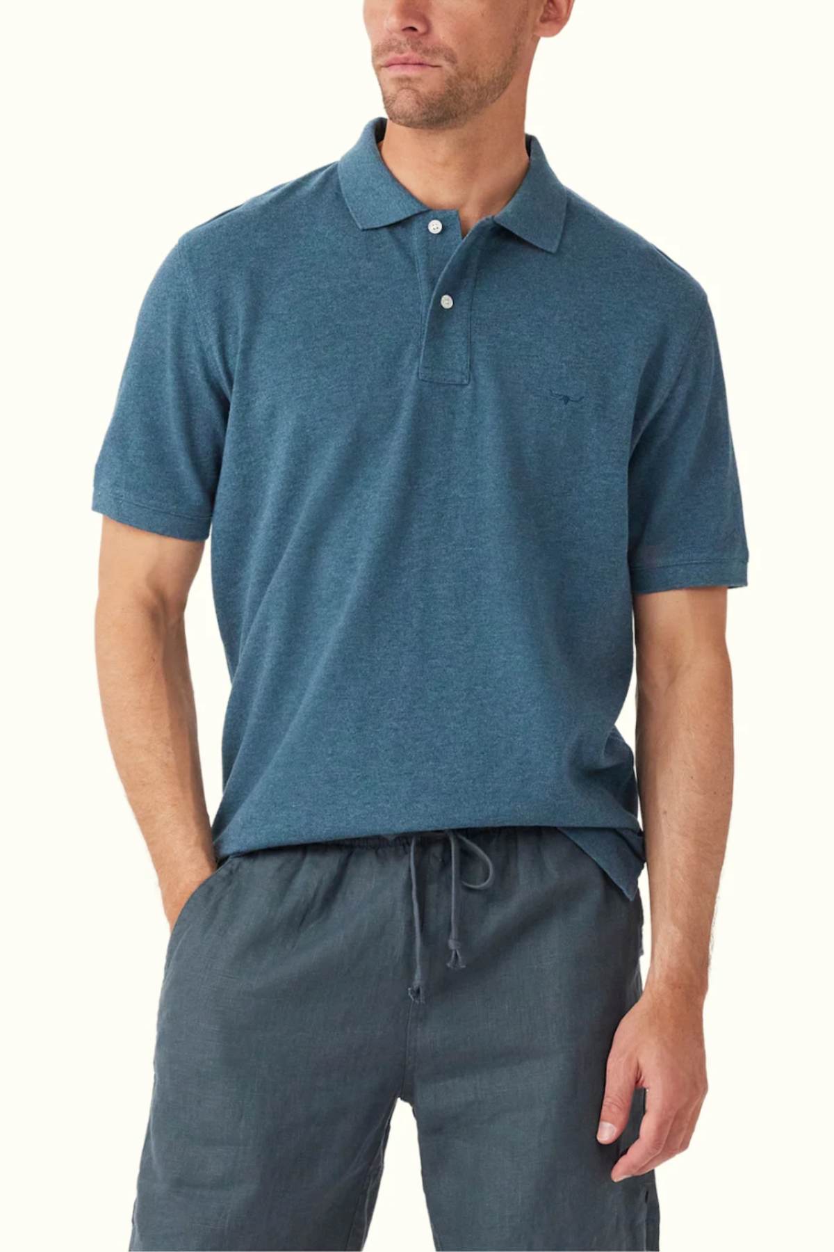 A man wearing a RM Williams Rod Polo in classic fit teal, crafted from organic cotton, alongside drawstring pants, stands with one hand in his pocket. The polo shirt features a collar and buttons against a plain background.