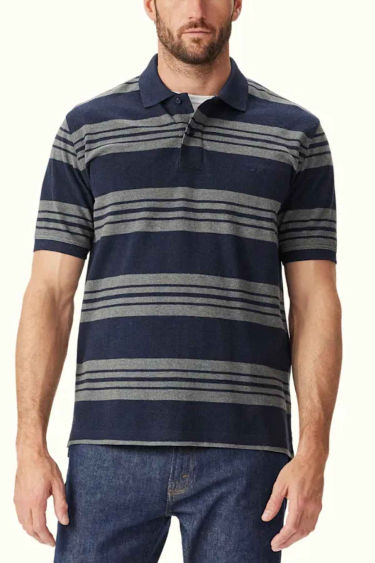 A man is wearing the RM Williams Rod Polo, a navy blue cotton shirt with horizontal gray stripes and Longhorn embroidery. The breathable cotton polo features short sleeves and a collar. He is also dressed in blue jeans, standing against a plain white background.