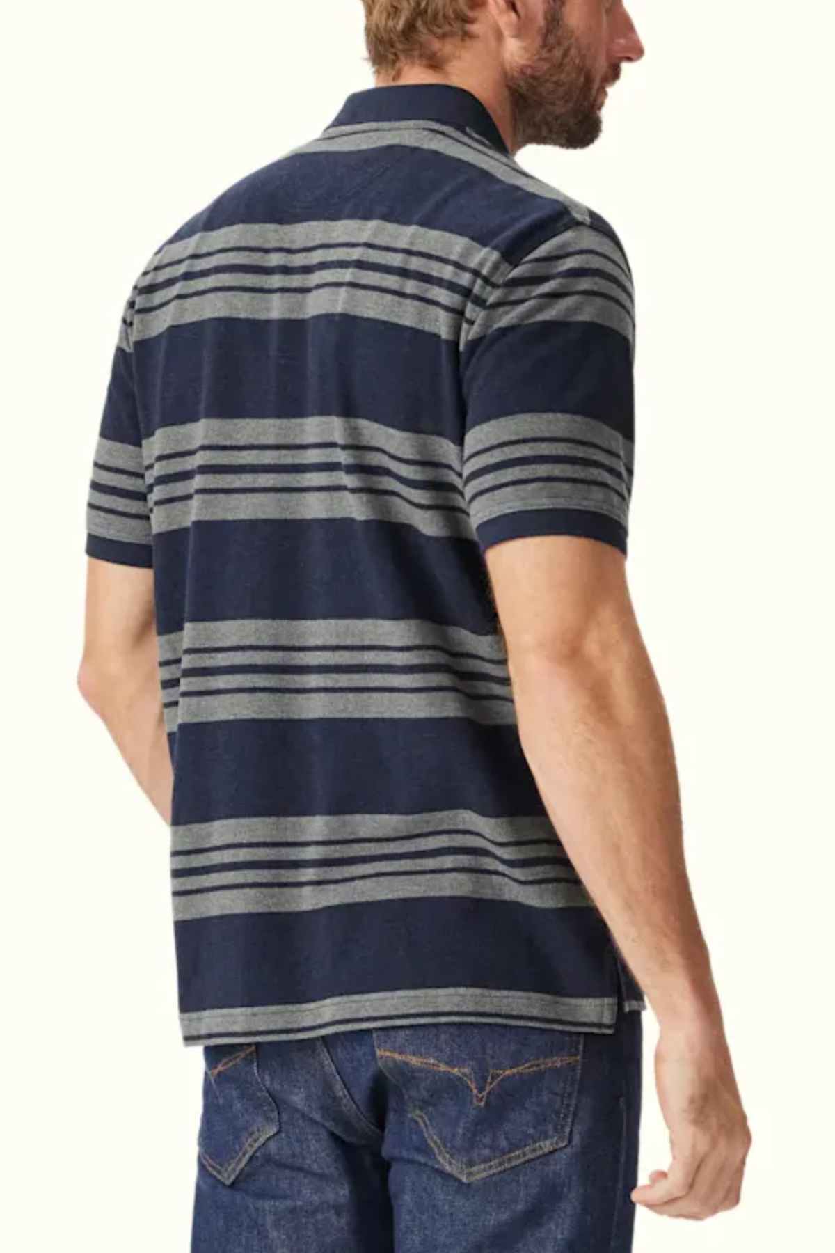 A man with a beard, seen from the back, is wearing an RM Williams Rod Polo in dark blue with gray horizontal stripes and blue jeans. The breathable cotton fabric ensures comfort as he stands against a plain, light-colored background.