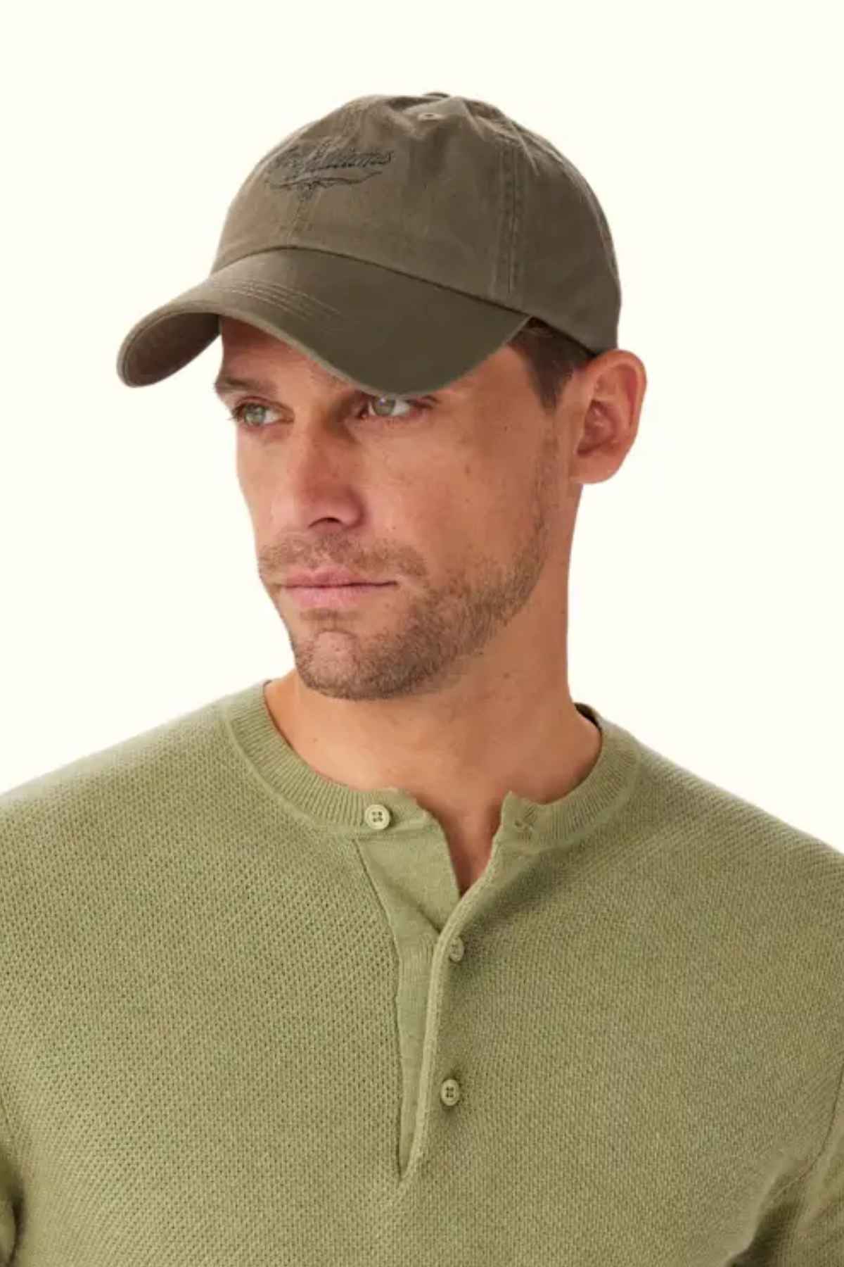 A person wearing the RM Williams Script Longhorn Cap in olive green, which features a heritage script logo, along with a matching textured henley shirt in organic cotton, looks to the side against a neutral background.