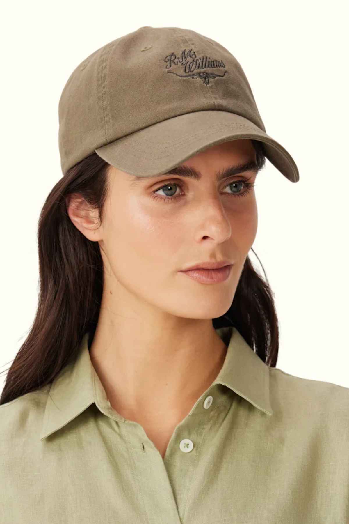 A person wearing a RM Williams Script Longhorn Cap in olive green, crafted from washed cotton twill, pairs it with a matching button-up shirt. The cap features a heritage script logo with text against a plain, light-colored background.
