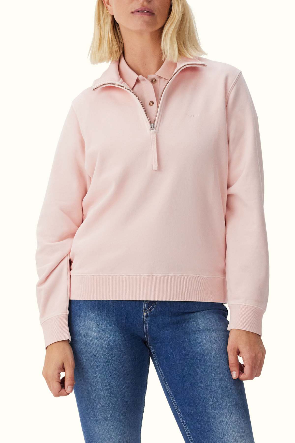 A person with shoulder-length blonde hair is wearing an RM Williams Trickett light pink quarter-zip sweatshirt over a buttoned pink collared shirt. They are also wearing blue jeans.