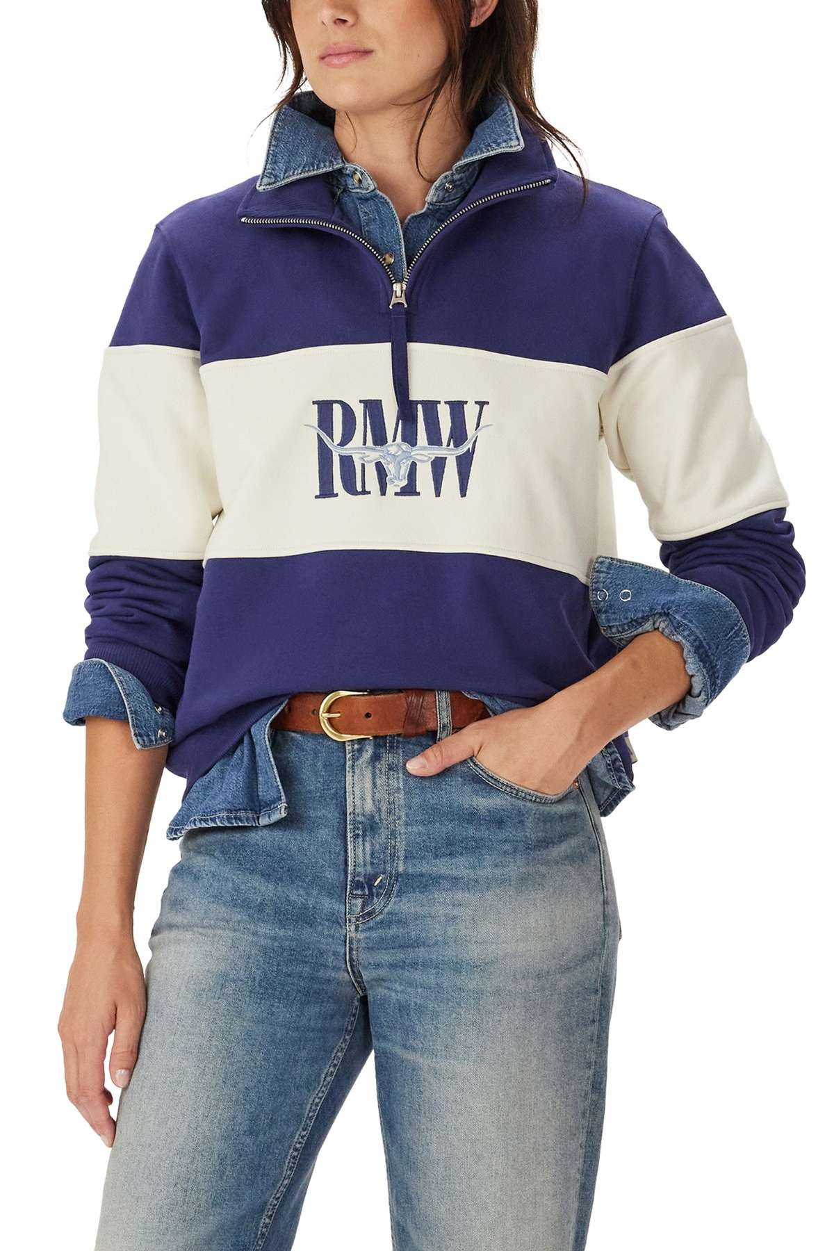 A woman wears light blue jeans, a blue denim shirt, and the RM Williams Trickett Panel Sweatshirt—a blue and white quarter-zip pullover with "RMW" on the front. Her hands are relaxed, and brown hair partially covers her face.