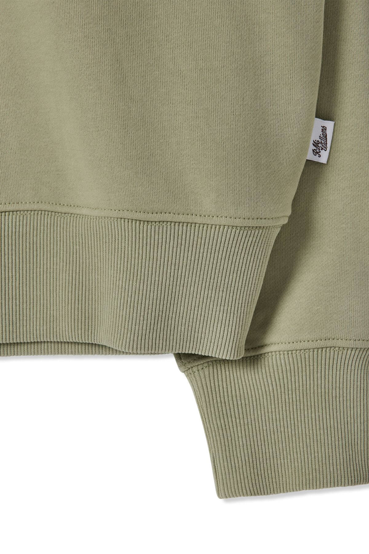 Close-up of the RM Williams Trickett Zip Neck Sweatshirt in light green, showcasing ribbed cuffs and hem. A small branded fabric tag is attached to the side seam. The cotton terry fabric texture and stitching details, with subtle longhorn embroidery, are also visible.