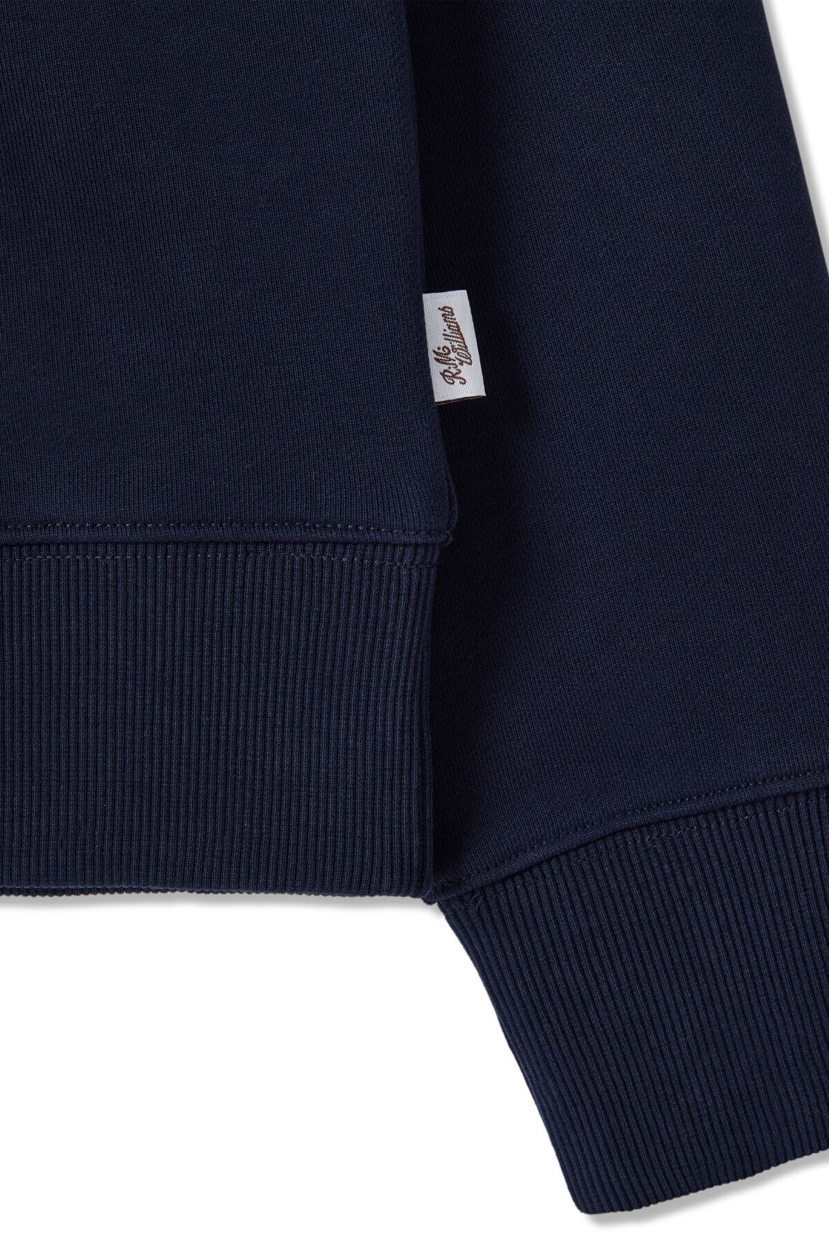 Close-up of the RM Williams Trickett Zip Neck Sweatshirt’s ribbed hem and cuff in navy blue cotton terry, featuring a small white fabric tag with cursive writing sewn into the seam.