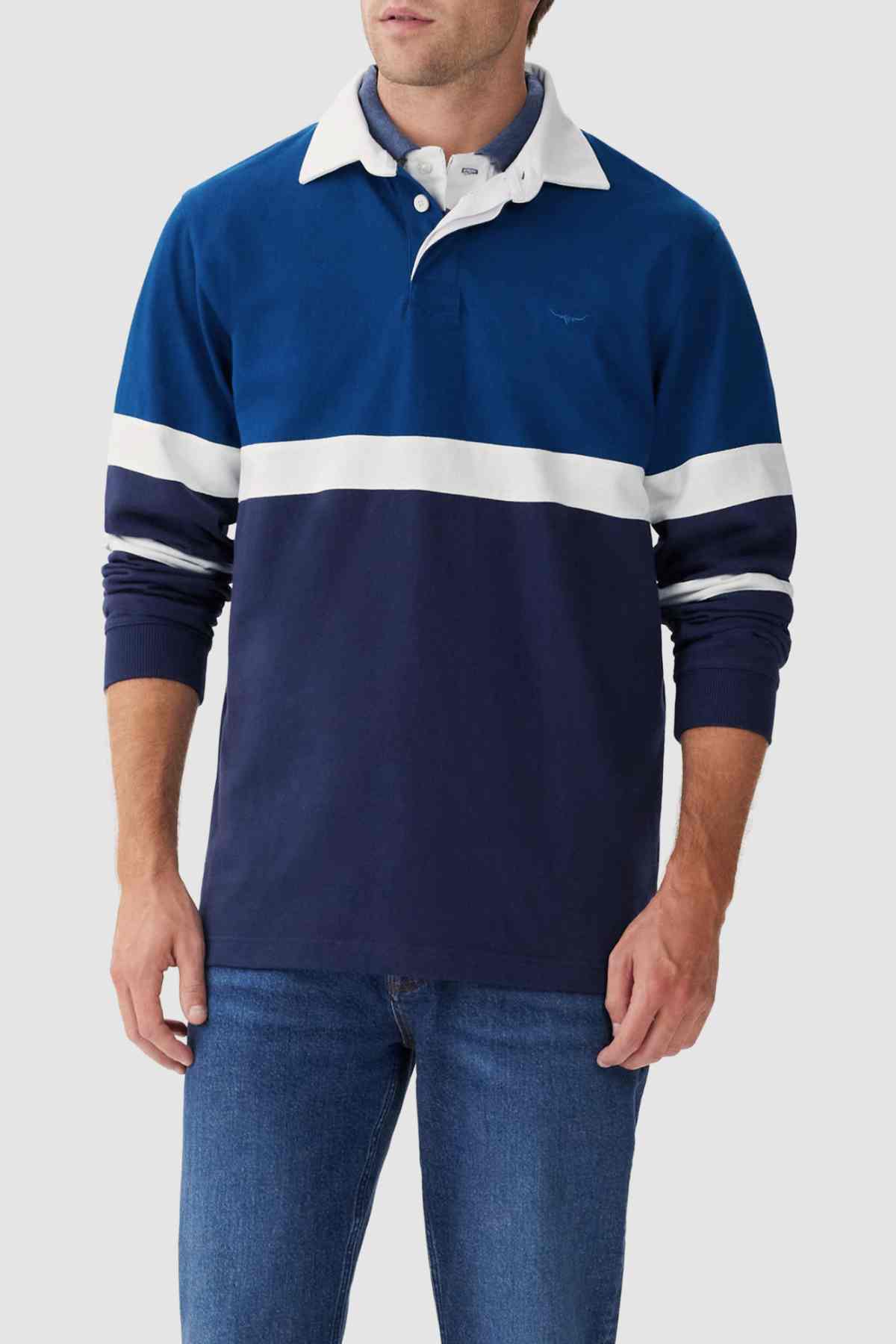 A person wearing the RM Williams Tweedale Rugby, a heavyweight blue rugby shirt featuring longhorn embroidery on the chest and a white collar. The person is also wearing blue jeans, set against a plain and light-colored background.