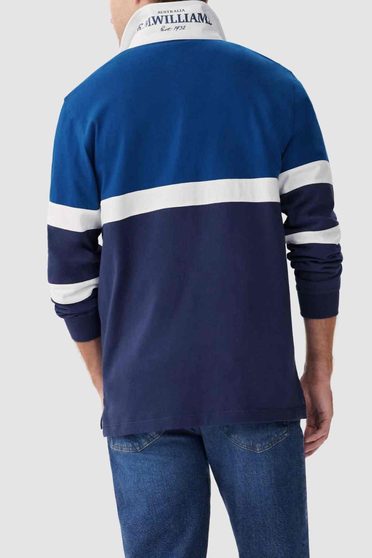 A person is seen from the back wearing an RM Williams Tweedale Rugby shirt, characterized by its blue and white long sleeves and a horizontal white stripe across the chest and upper arms. The collar, featuring text, is contrasted in white. Complementing the outfit, they are also sporting blue jeans.