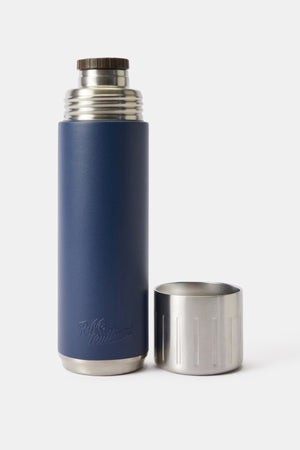 The RM Williams Vacuum Flask, featuring double-wall stainless steel insulation and a navy blue body, stands upright with its metallic screw-on lid placed upside down on a plain white background.