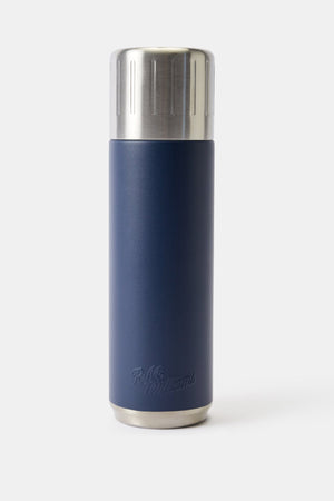 The RM Williams Vacuum Flask, in navy blue with double wall insulation and a stainless steel lid, stands upright on a plain light background. A discreet embossed logo is featured near its base.