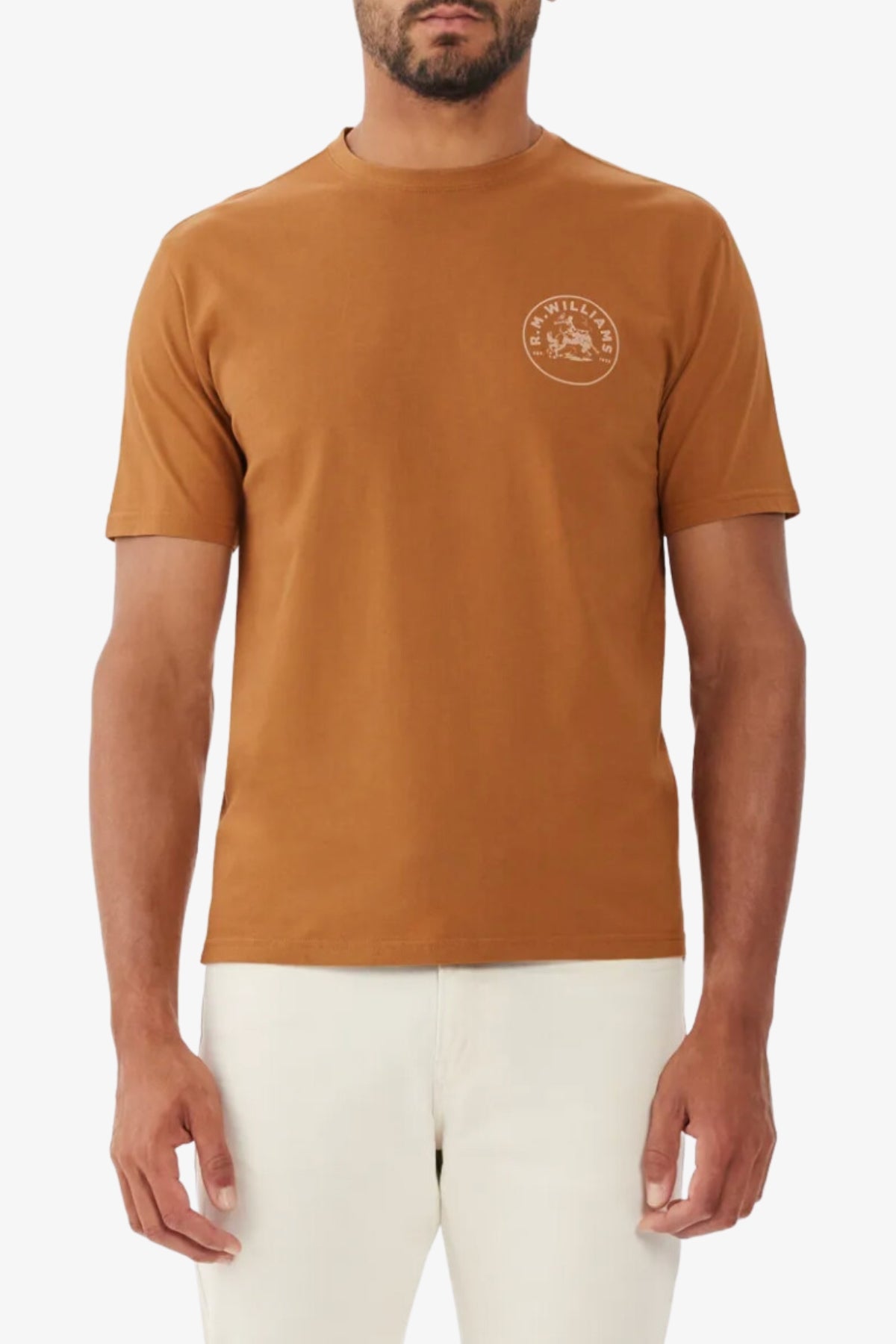 Someone is wearing a brown RM Williams Wondai T-Shirt, made from lightweight cotton jersey with a white circular logo on the left chest, paired with light-colored pants against a plain white background.