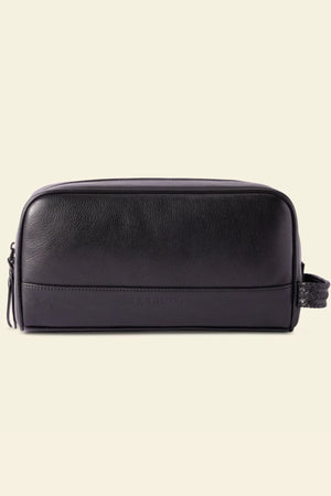 The RM Williams Farrier Signature Washbag is a black tumbled grain leather toiletry bag with a rectangular design, zip closure, water-resistant lining, and woven side handle, shown on a plain cream background.
