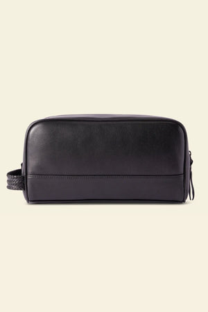 The RM Williams Farrier Signature Washbag is a rectangular black tumbled grain leather toiletry bag with a minimalist design, woven side handle, zipper closure, and water-resistant lining, shown on a light beige background.