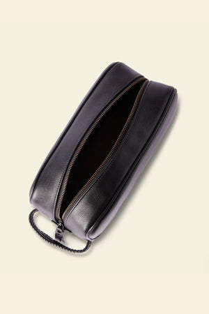 The RM Williams Farrier Signature Washbag, crafted from black tumbled grain leather, features a textured handle, water-resistant lining, and a zippered top. Shown against a light beige background with its empty interior visible.