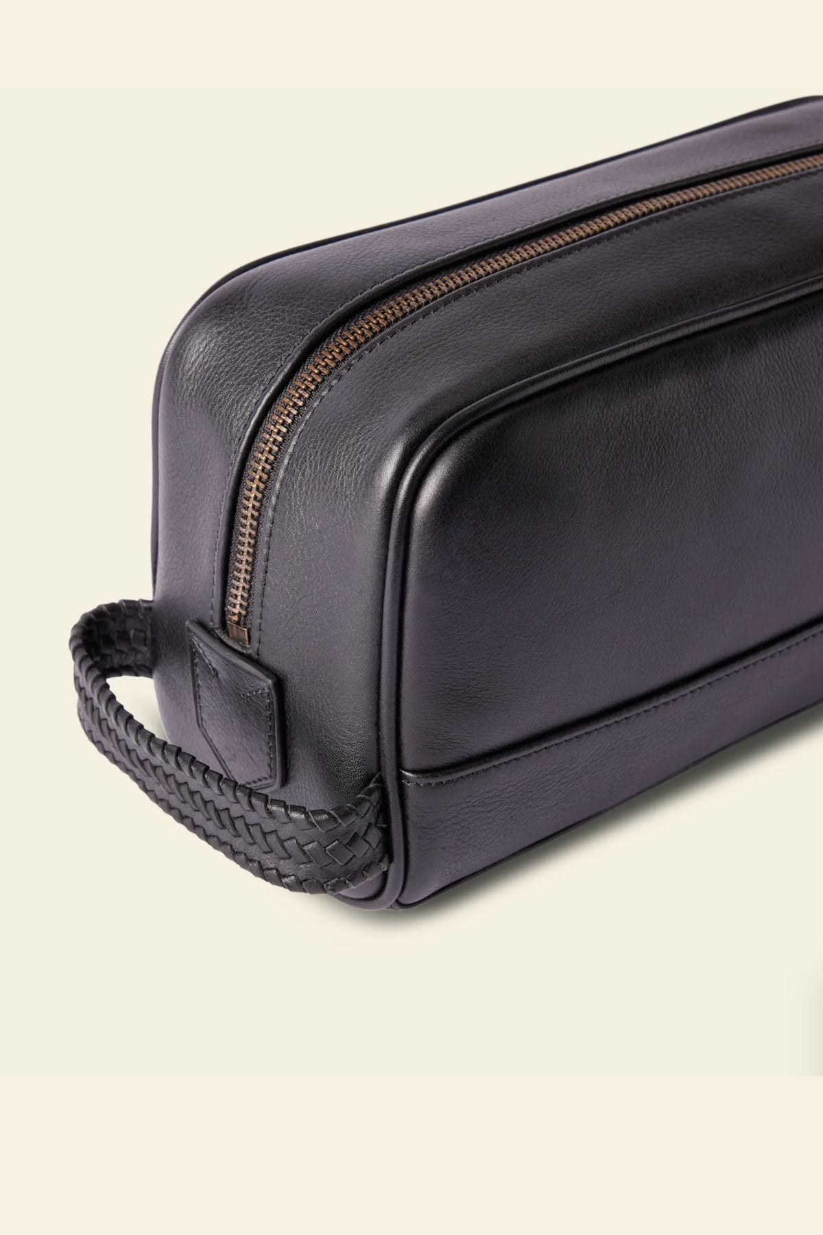 The RM Williams Farrier Signature Washbag is a black tumbled grain leather toiletry bag with a gold zipper, woven handle, and water-resistant lining, showcased on a light beige background.