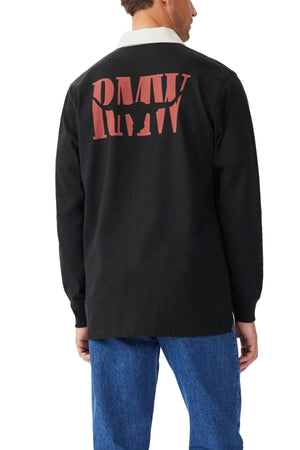 A person, turned away from the camera, wears the RM Williams Branded Rugby—a black heavyweight brushed cotton shirt with full-length sleeves and bold red "RMW" embroidery on the back—paired with blue jeans.
