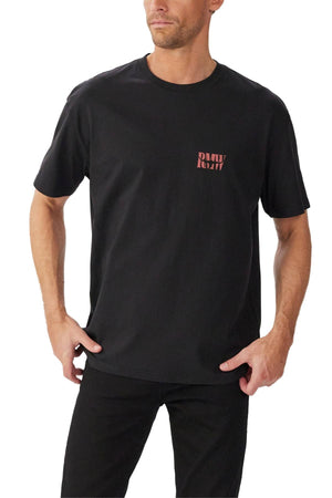 A man wears the RM Williams Branded T-Shirt, a relaxed fit black tee with short sleeves and small red "PAHU RUAH" lettering on the left chest, paired with black pants. He stands facing slightly to the side, hands near his pockets.