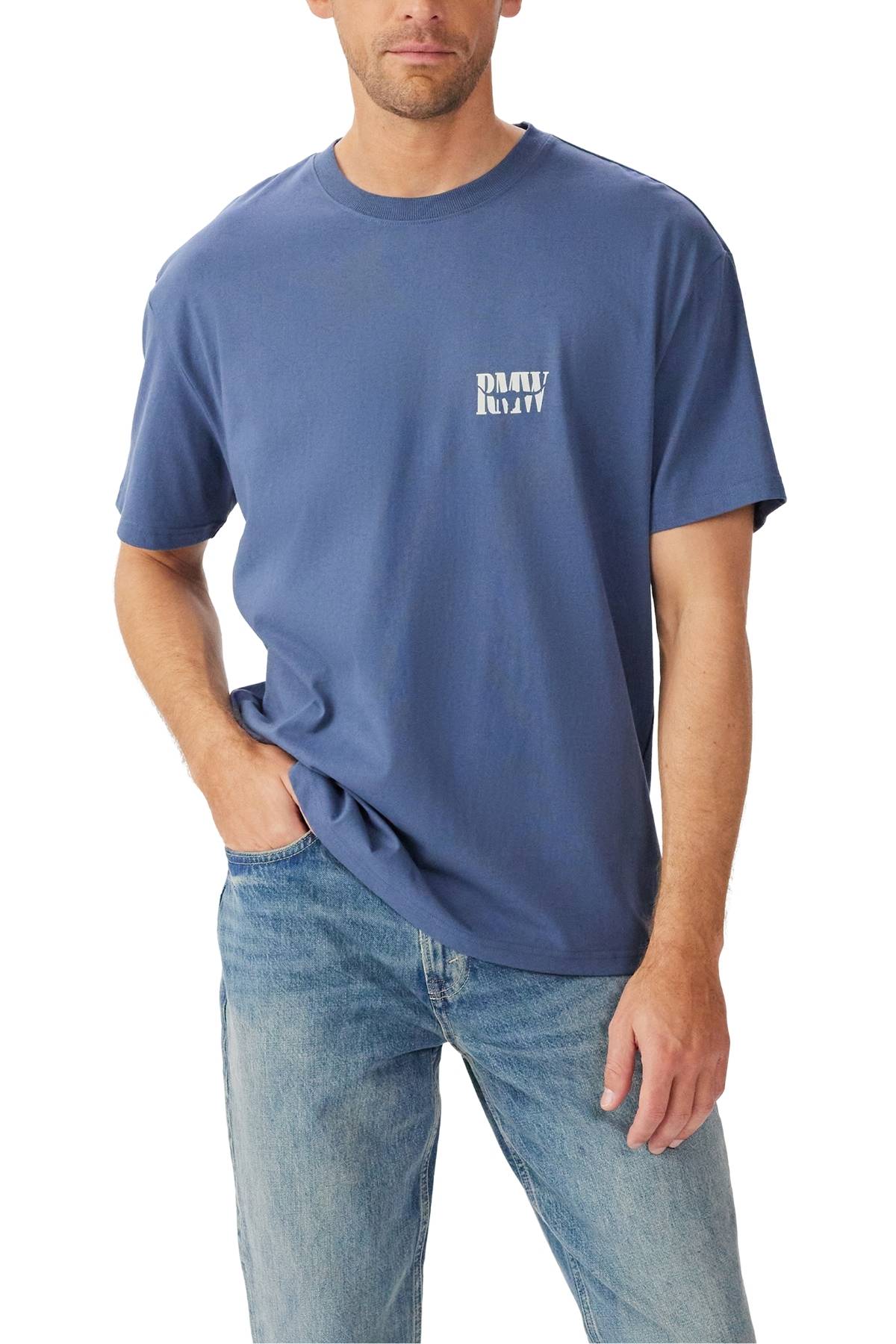 A man wears the RM Williams Branded T-Shirt in blue with a small heritage logo on the chest and light blue jeans, posing with one hand in his pocket.