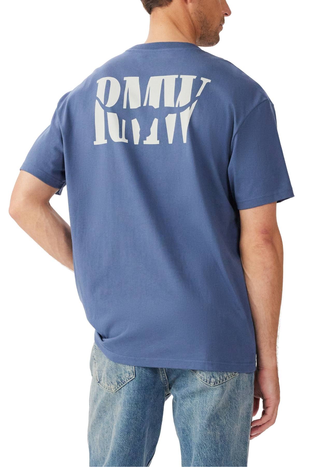 A man wears the RM Williams Branded T-Shirt, showcasing a large white "RMV" heritage logo on the back. He stands with one hand on his hip, facing away from the camera, and pairs it with light blue jeans for a casual look.