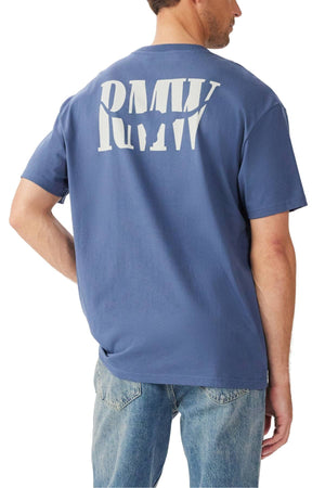 A man wears the RM Williams Branded T-Shirt, showcasing a large white "RMV" heritage logo on the back. He stands with one hand on his hip, facing away from the camera, and pairs it with light blue jeans for a casual look.
