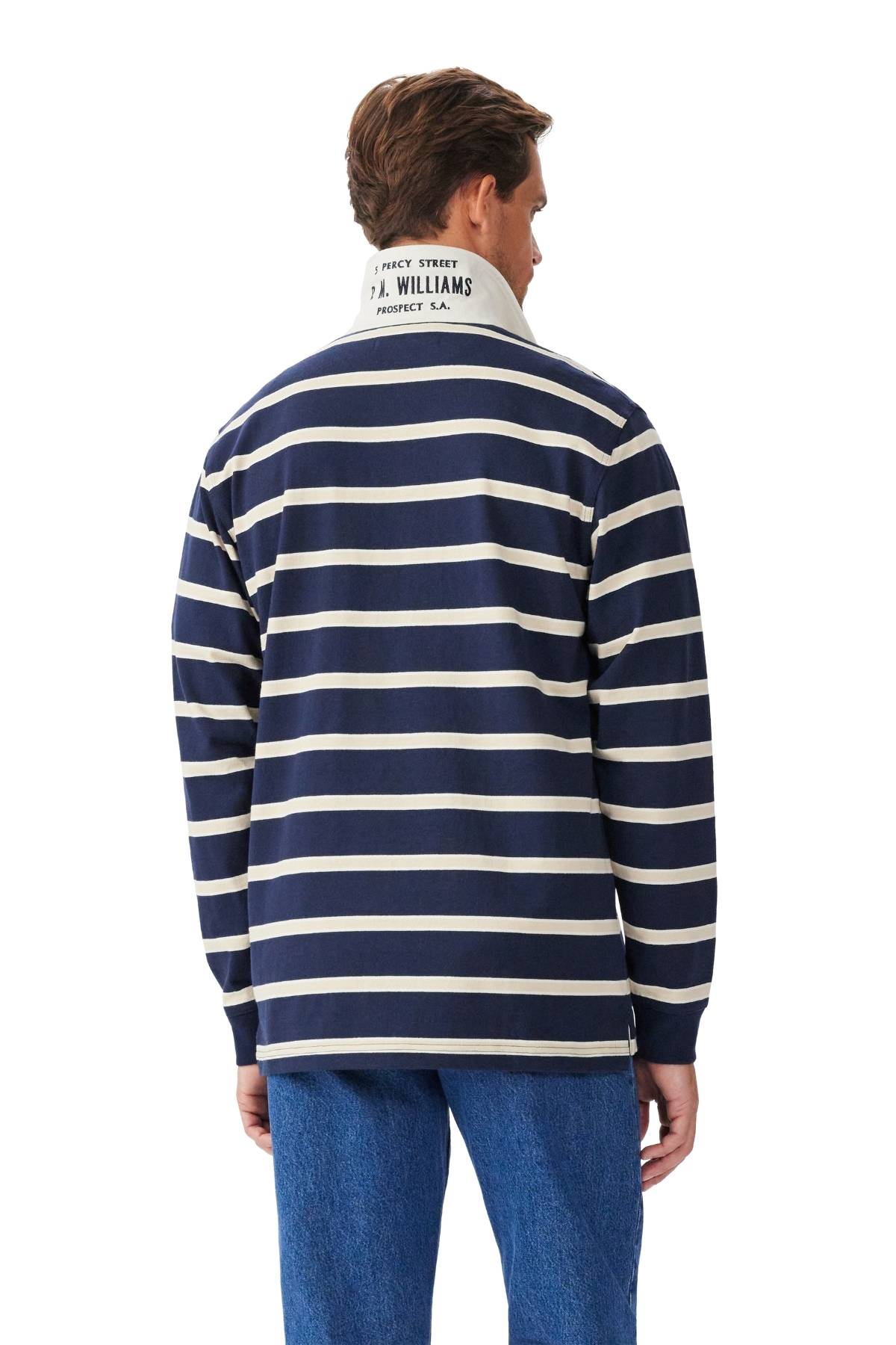 A man is shown from the back wearing the RM Williams Brunswick Rugby—a navy blue and white striped long-sleeve shirt with a twill collar featuring text—paired with blue jeans.
