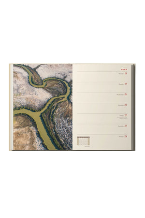 An open RM Williams Outback Diary 2024 shows a weekly calendar (March 18–24) on the right and an evocative photo of a winding green river through snowy, brown terrain on the left.