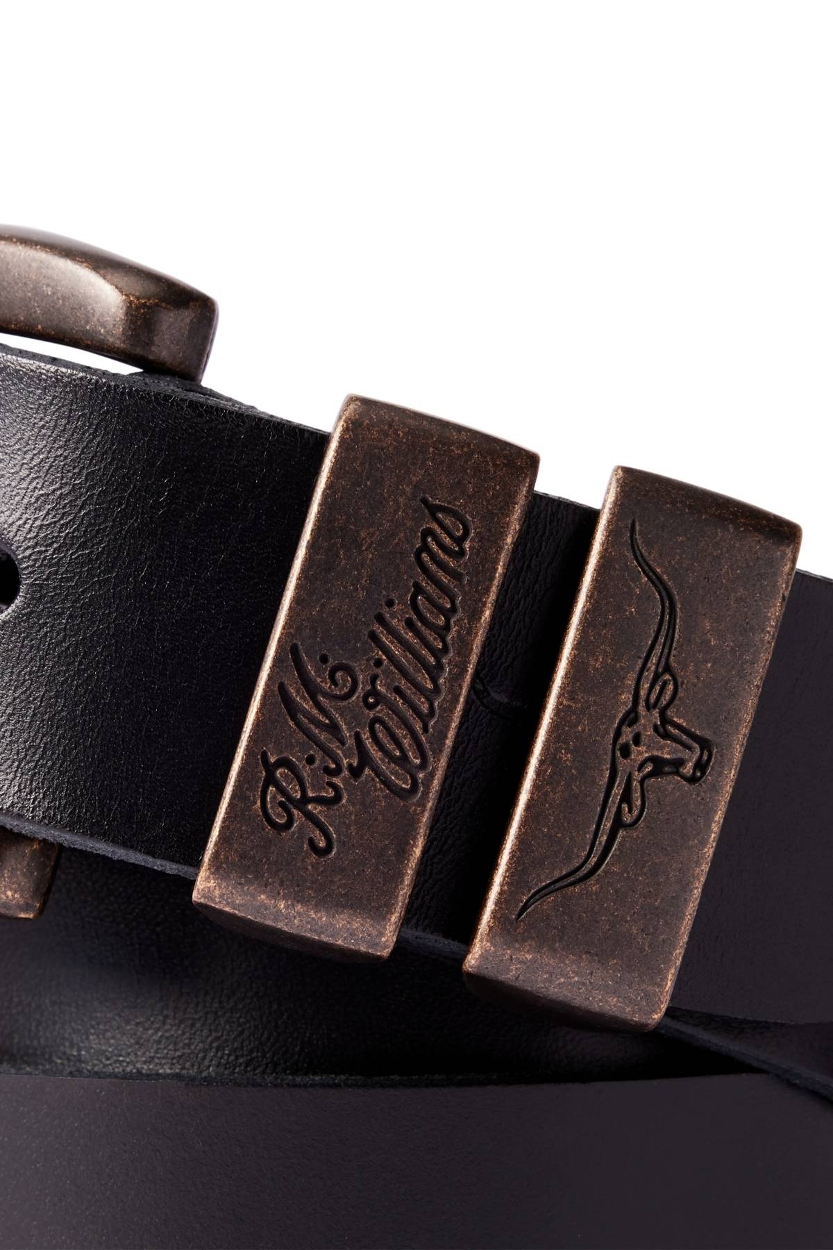 A close-up of the RM Williams Drover Belt in black pull-up leather, featuring antique brass buckles—one engraved with "R.M.Williams," the other with a longhorn steer logo—showcasing its classic double keepers design.
