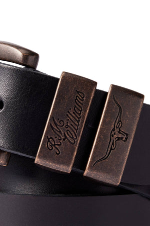 A close-up of the RM Williams Drover Belt in black pull-up leather, featuring antique brass buckles—one engraved with "R.M.Williams," the other with a longhorn steer logo—showcasing its classic double keepers design.