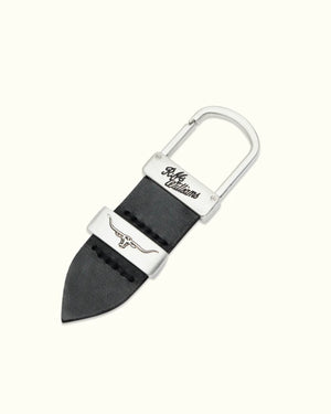 The RM Williams Drover Key Fob boasts a silver keychain with a black leather strap, adorned with antique brass hardware and a metal bull's head engraving. A metal piece featuring text on the top adds to its stylish functionality.