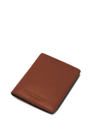 The RM Williams Farrier Fold Cardholder is a slim, brown full grain leather wallet with visible stitching, shown closed at a slight angle on a white background. The embossed R.M.Williams logo appears near the bottom left corner.