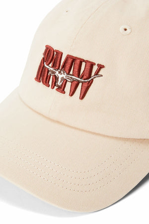 The RM Williams Heritage Twill Cap is a beige organic cotton cap with "RAW" embroidered in bold red, a brown and white steer skull above the text, and an adjustable strap for a custom fit.