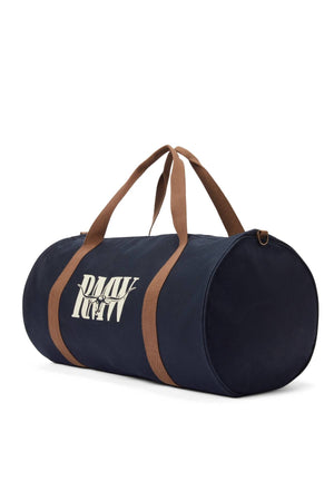 The RM Williams Heritage Bag is a navy blue cylindrical cotton canvas duffel featuring tan straps, an adjustable crossbody strap, and a white heritage logo printed on the side, shown against a plain white background.