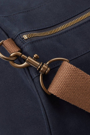 Close-up of the RM Williams Heritage Bag in navy blue cotton canvas, featuring a brass zipper and clasp, and a brown woven adjustable crossbody strap attached to metal hardware.