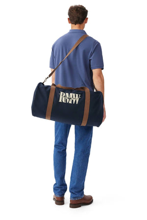 A man in a blue polo, jeans, and brown shoes stands facing away, carrying the RM Williams Heritage Bag—navy cotton canvas with an adjustable crossbody strap and white heritage logo—over his shoulder.