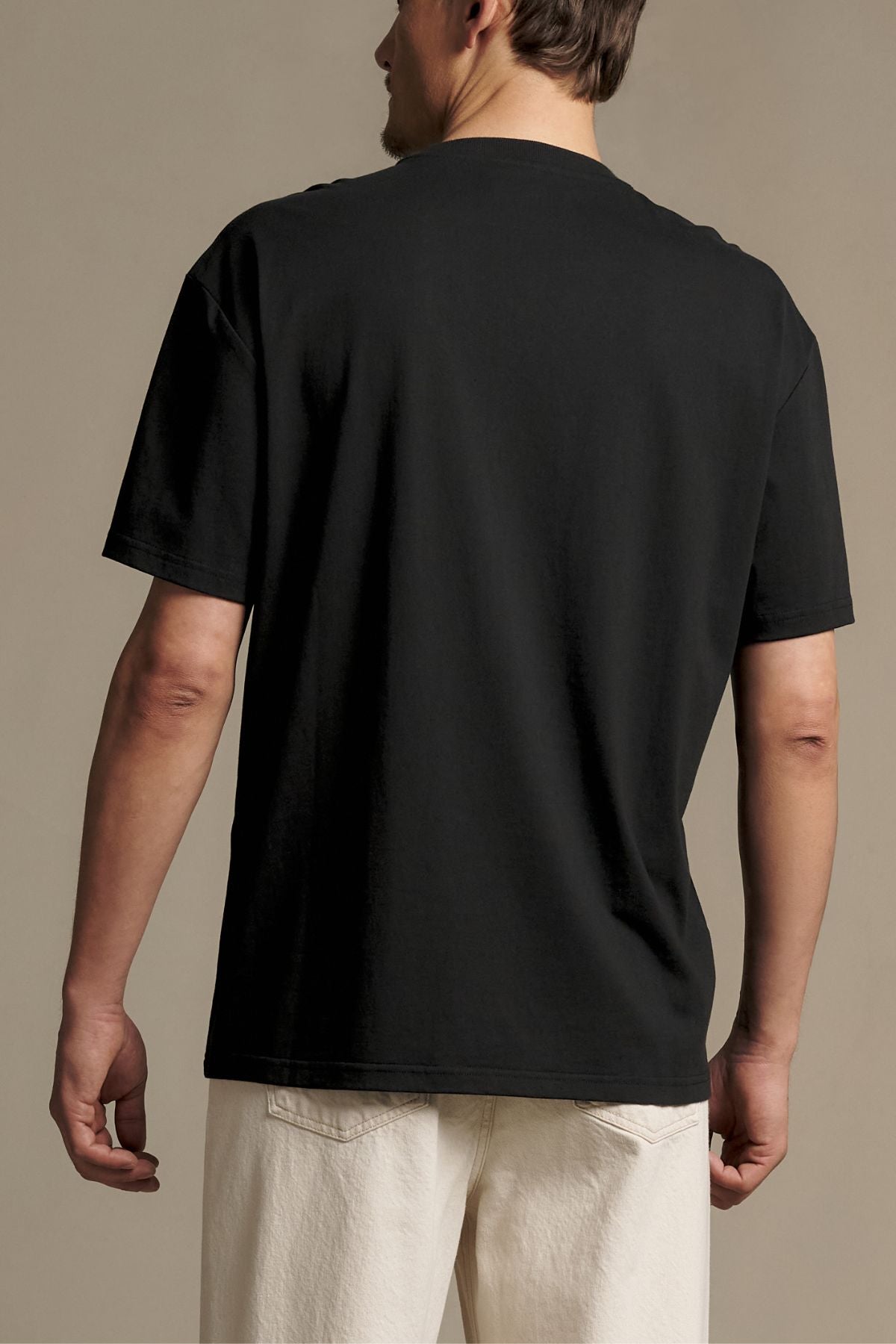 A person stands facing away, wearing the RM Williams Holt Tee in black—a relaxed fit t-shirt with a ribbed crew neck—paired with light-colored pants against a neutral background.