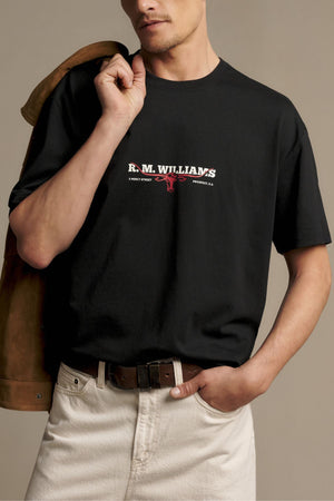 Wearing the RM Williams Holt Tee—a relaxed fit, black t-shirt crafted from soft Australian cotton and featuring a red logo—paired with off-white pants, this person holds a brown jacket over their shoulder while standing against a neutral background.