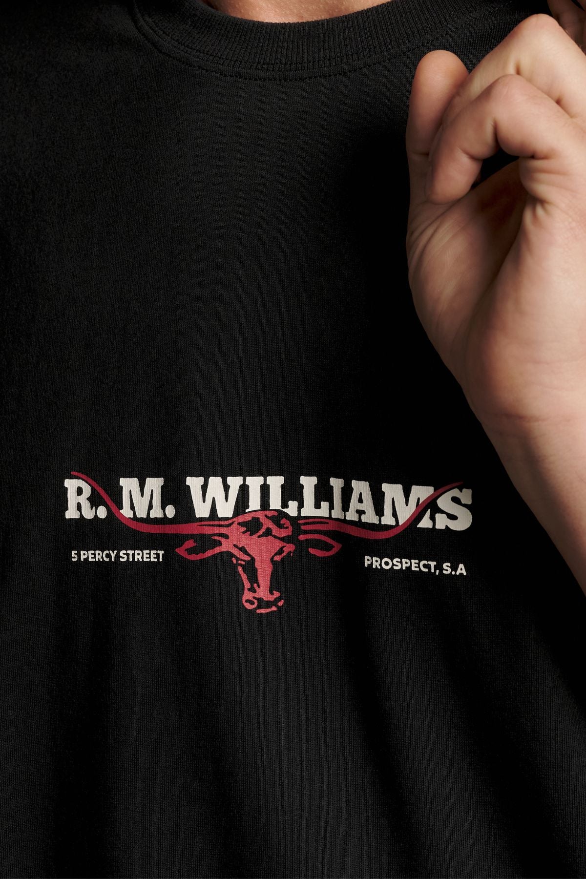 A person is wearing the RM Williams Holt Tee, a black relaxed fit shirt featuring “R.M. Williams” and a red bull logo, along with “5 Percy Street” and “Prospect, S.A.” printed on it. The tee’s ribbed crew neck and premium Australian cotton enhance its comfortable style. A hand is partially visible near the shirt.
