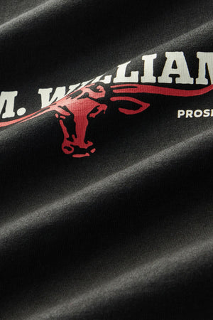 Close-up of the black RM Williams Holt Tee made from Australian cotton, featuring bold white text partially displaying “M. WILLIAM” and a red outline of a bull’s head. Additional smaller white text is visible on the right side, and the ribbed crew neck highlights the relaxed fit design of the t-shirt.